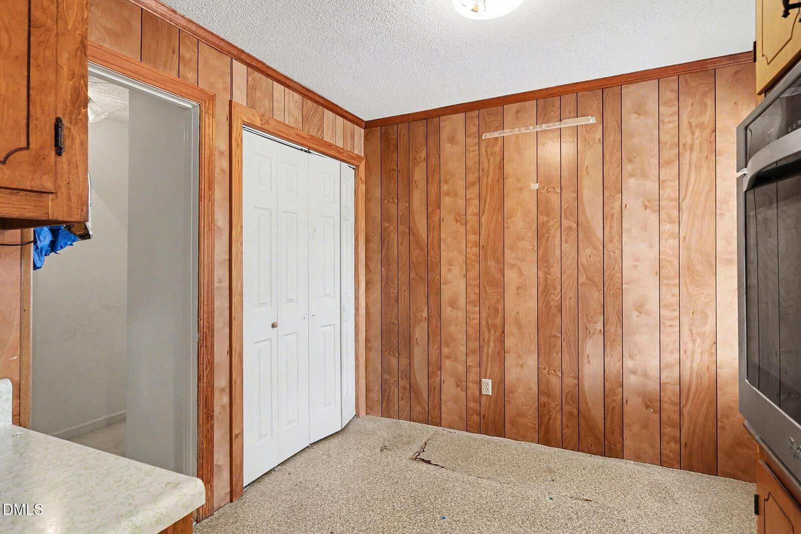 A small room with wood-paneled walls, double white closet doors, carpeted floor, and part of a kitchen countertop and oven visible. The room appears empty and has some wear on the carpet.