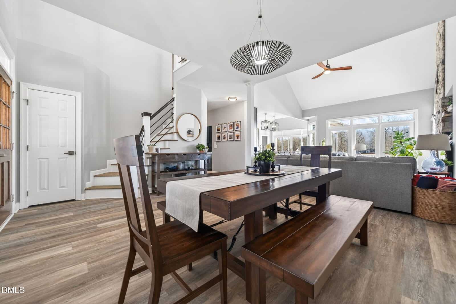 A modern open-concept dining and living area with wooden dining table and benches, grey sectional sofa, large windows, light wood flooring, and a staircase leading upstairs. Natural light fills the space.