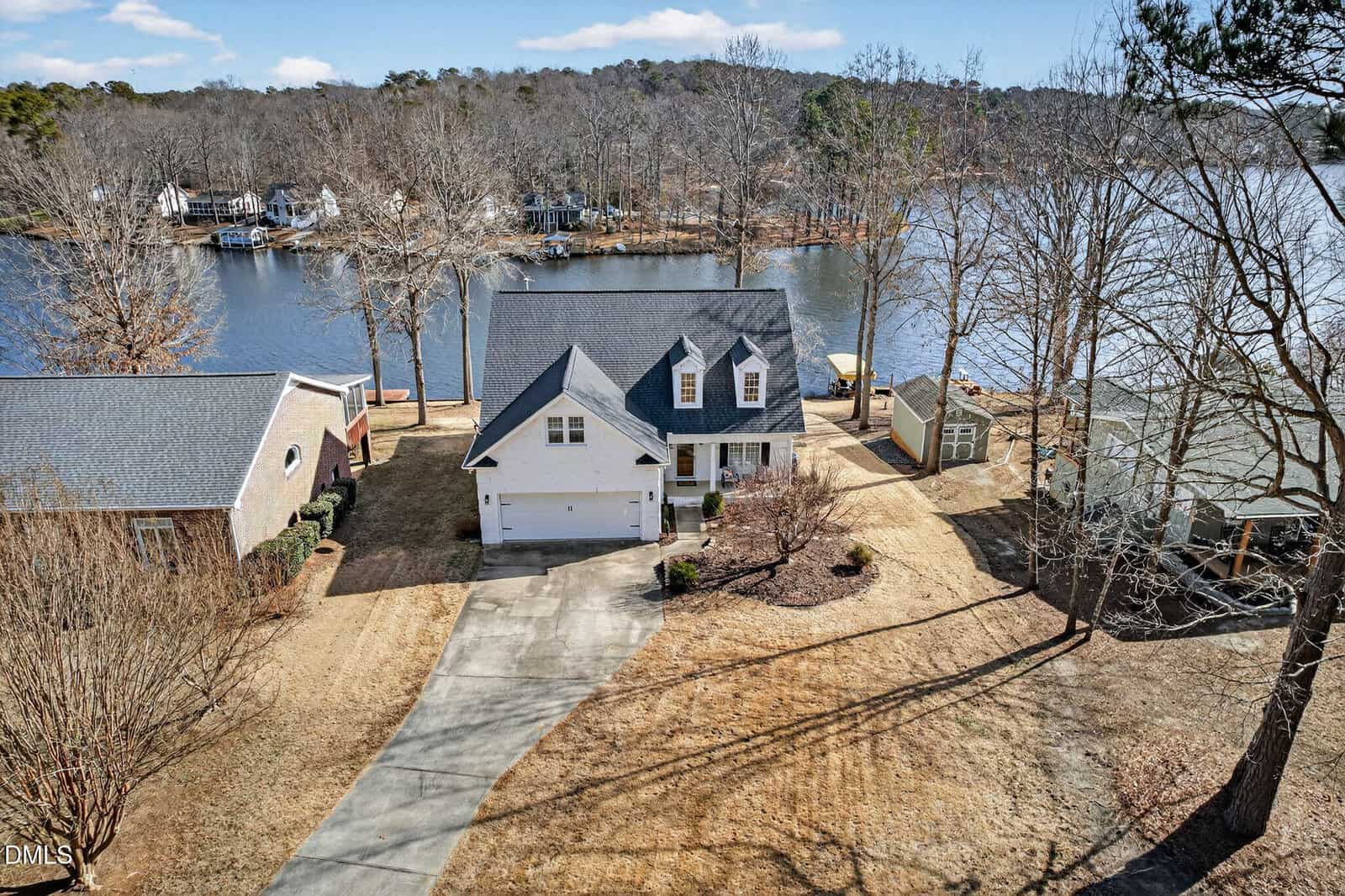 Aerial view of a two-story house with a white exterior and attached garage, situated by a lake with several docks. The yard is surrounded by bare trees and neighboring houses, and a winding driveway leads to the home.