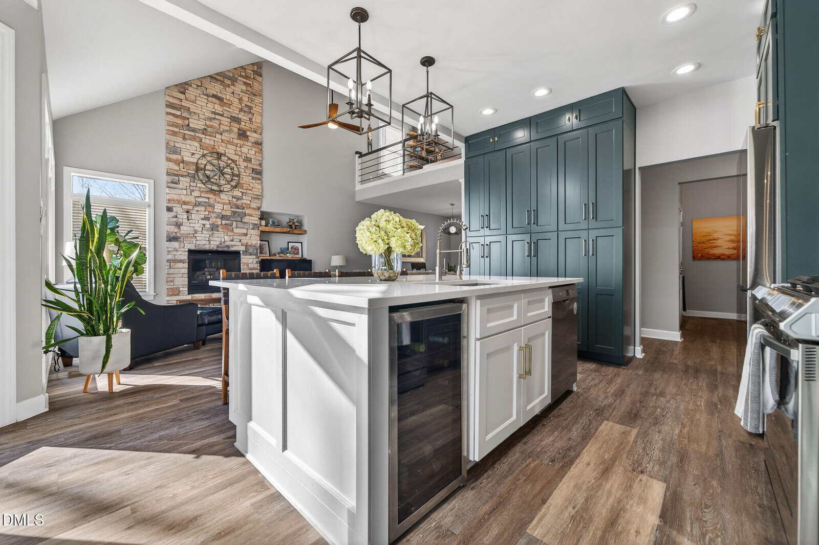 Modern kitchen with a large white island, dark green cabinets, wood floors, a fireplace with stone surround, and bright natural light. A bouquet of white flowers sits on the island.