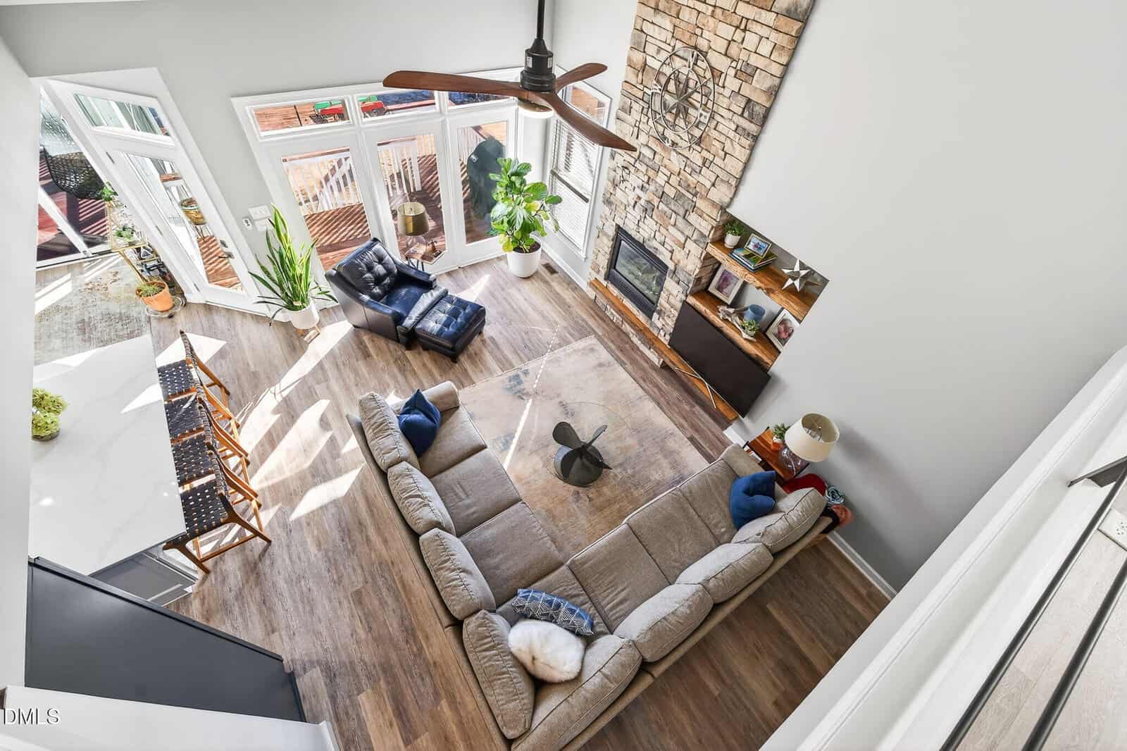 A bright, modern living room seen from above features a large sectional sofa, a stone fireplace, a wall clock, a coffee table, indoor plants, and sunlight streaming through tall windows overlooking a deck.
