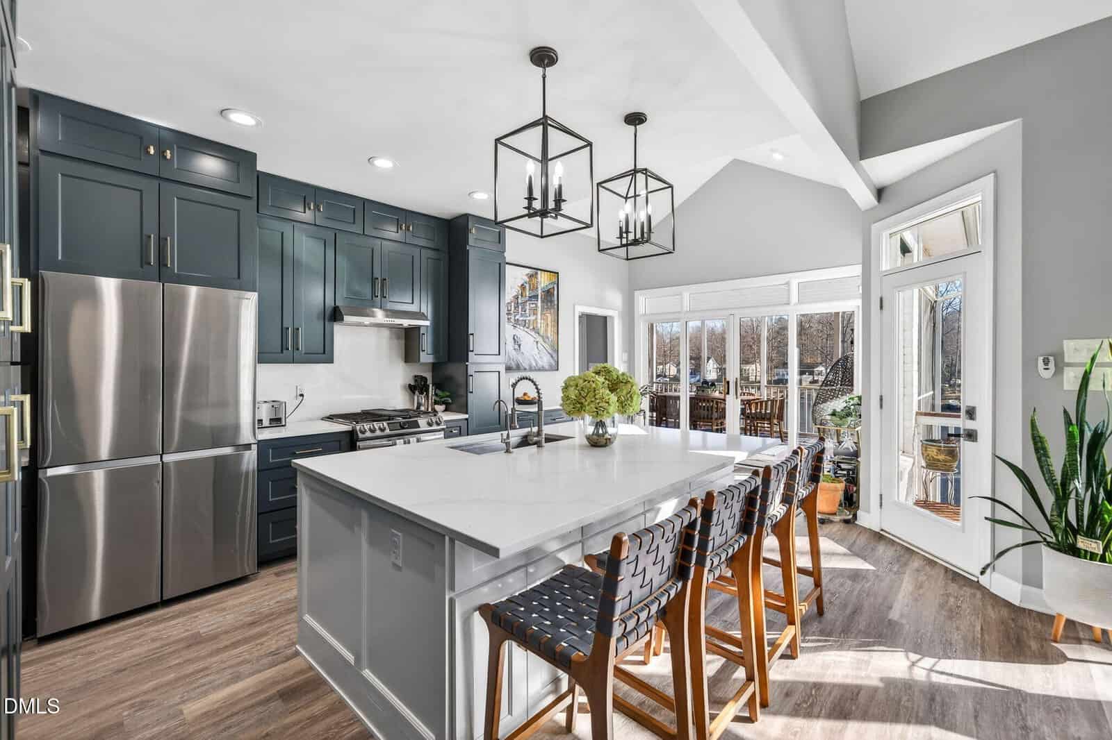 Modern kitchen with dark cabinets, stainless steel appliances, a white island with four woven barstools, hanging lights, houseplants, and large windows letting in natural light.
