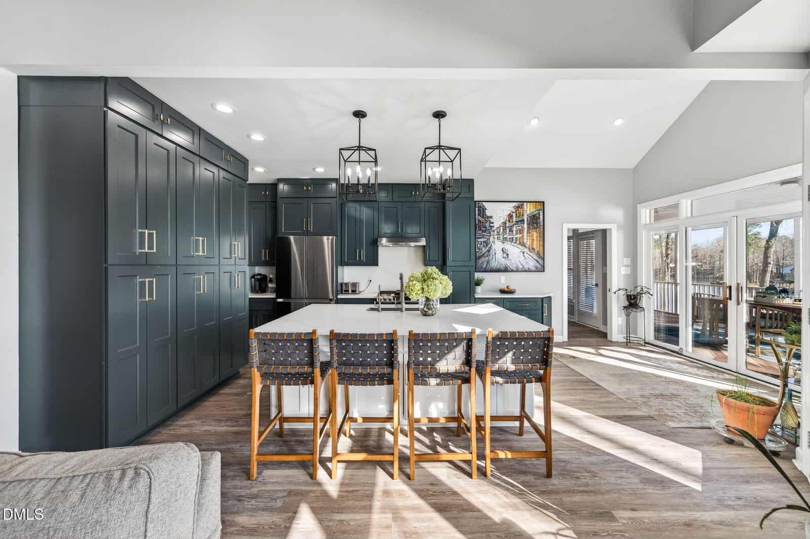 Modern kitchen with dark teal cabinets, stainless steel appliances, white island with four woven barstools, pendant lights above, wood floors, large windows, and a door leading to a sunny deck with outdoor seating.