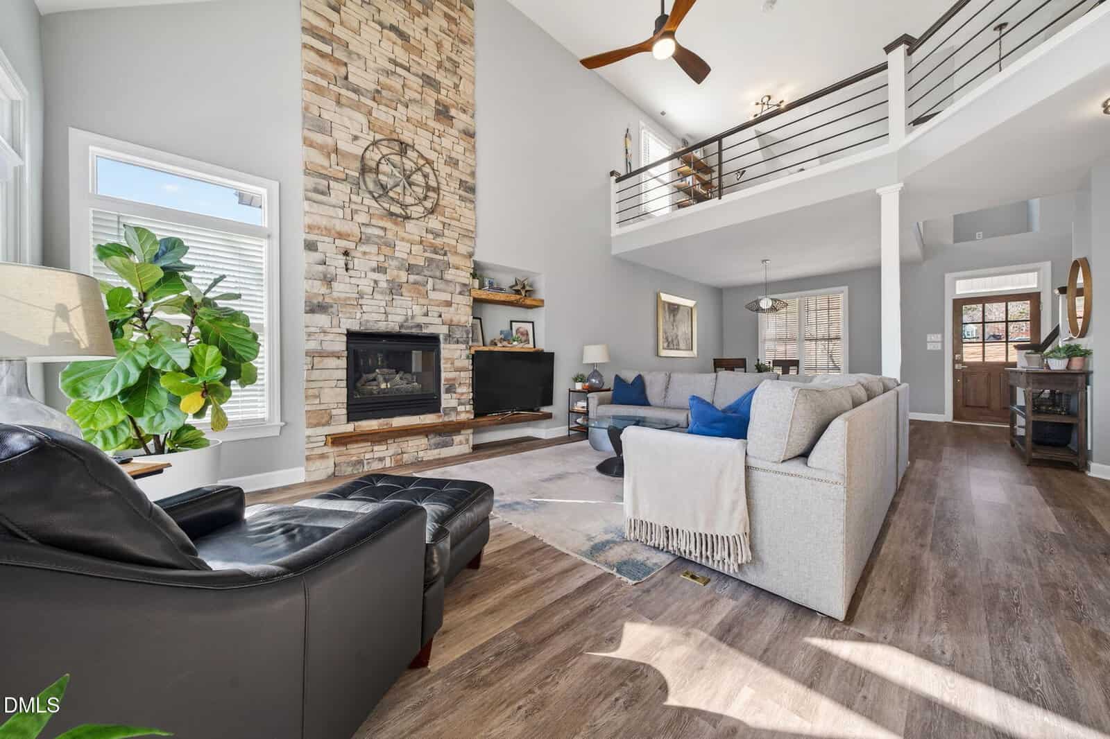 Bright living room with high ceilings, stone fireplace, modern furniture, wood floors, and a loft. Large windows let in natural light, and a plant sits by a black chair. TV and shelves are next to the fireplace.