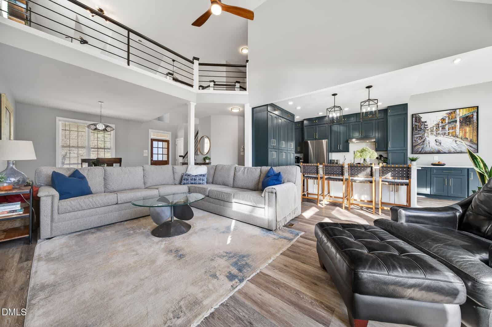 Open-concept living room with gray sectional, modern rug, black leather chair, and a kitchen in the background featuring blue cabinets, barstools, and pendant lights beneath a loft and ceiling fan.