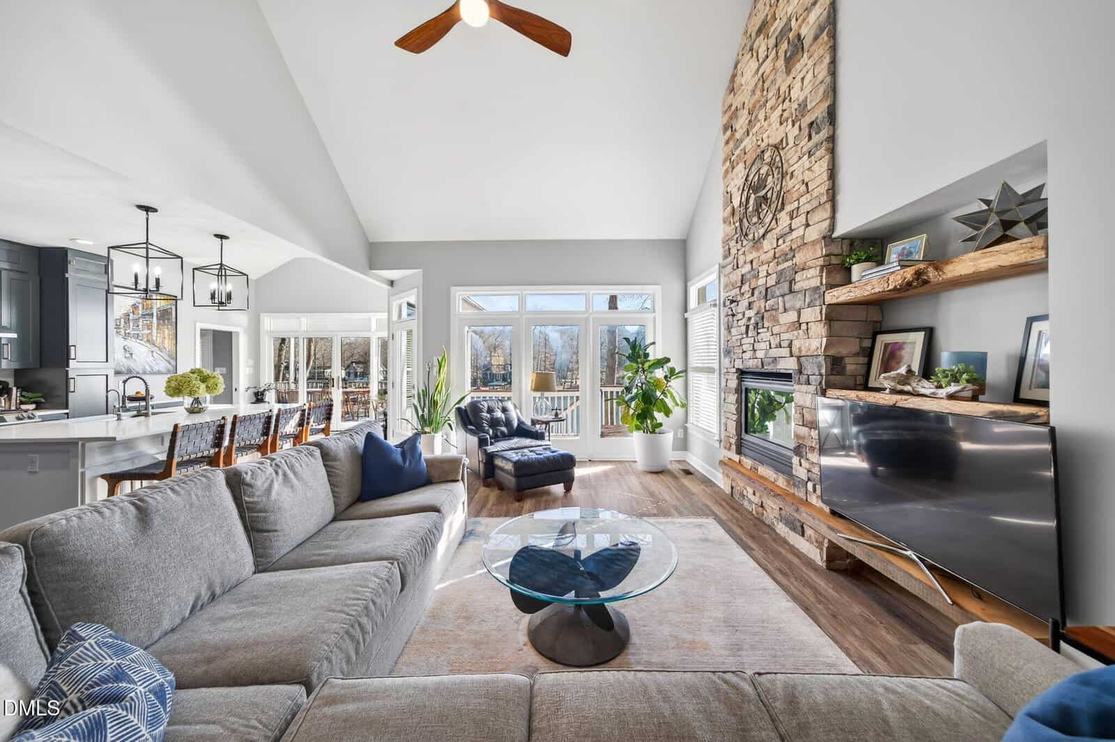 A bright, modern living room with a stone fireplace, mounted TV, gray sectional sofa, glass coffee table, and large windows overlooking a deck and trees. The space connects to an open kitchen and dining area.