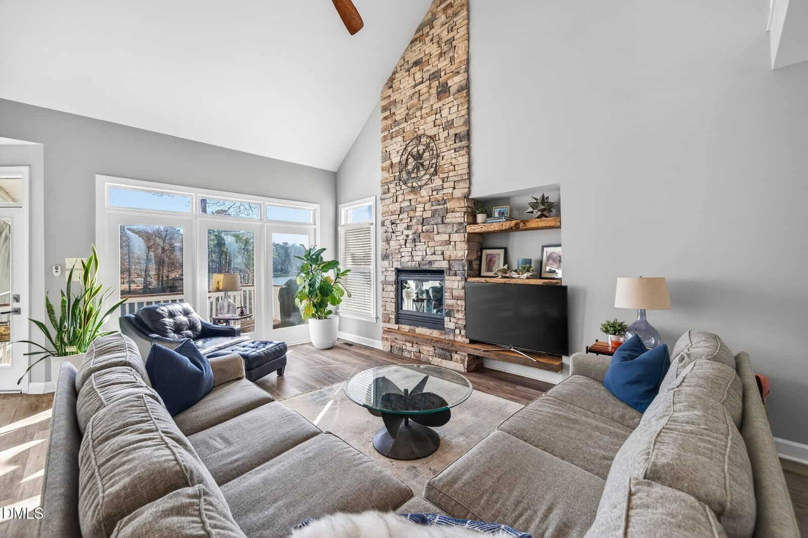 A bright living room with a high ceiling, stone fireplace, large windows, a sectional sofa, glass coffee table, plants, and a wall-mounted TV on a wooden console. Sunlight streams in from the windows.
