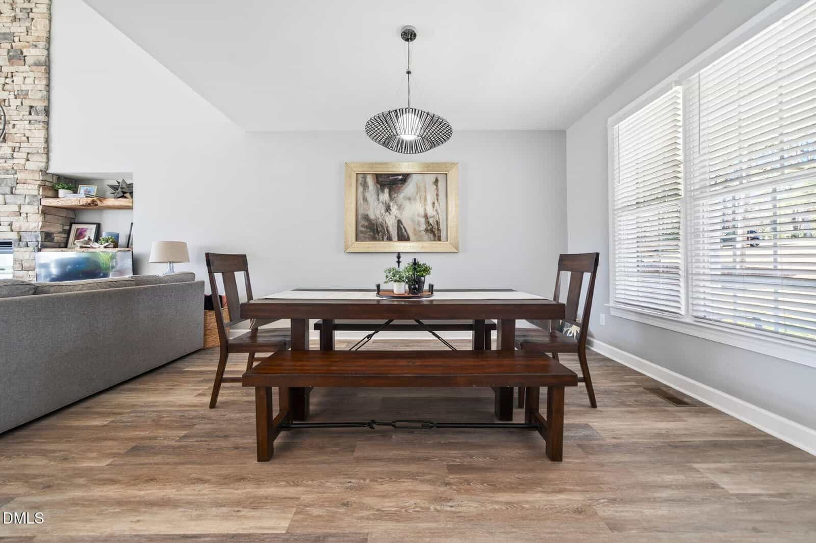A modern dining room with a wooden table, two chairs, and a bench. A pendant light hangs above the table, a painting is on the wall, and large windows with blinds let in natural light. A living area is partially visible on the left.