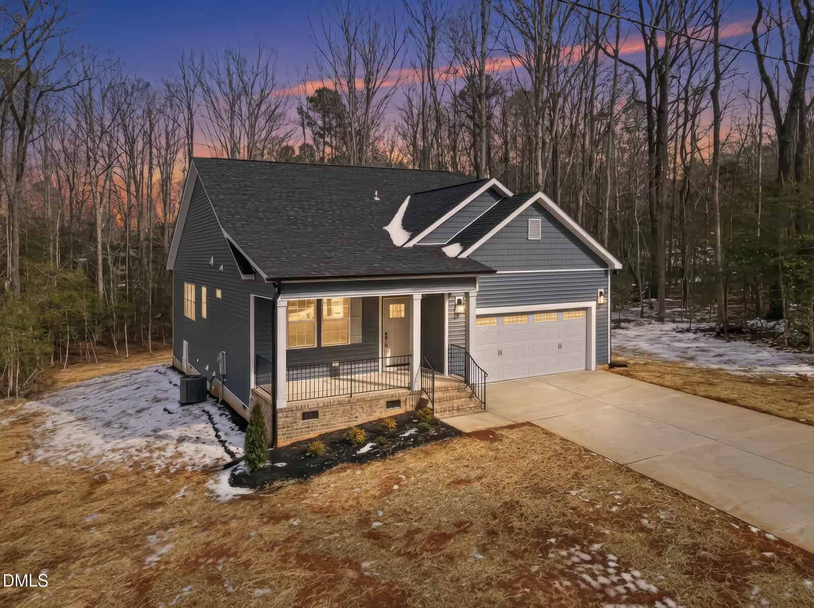 A modern gray house with a covered front porch and attached two-car garage, set on a spacious lot with a concrete driveway and surrounded by trees at sunset.