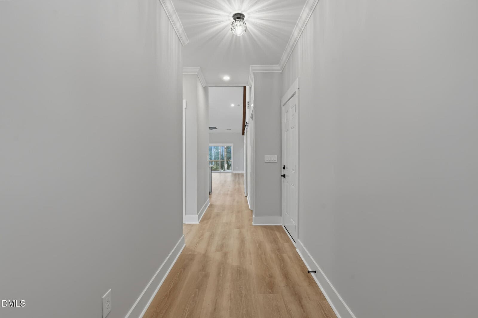 A modern hallway with light gray walls, crown molding, and light wood flooring, leading to a bright, open living area with large windows at the end. A white door is visible on the right side.