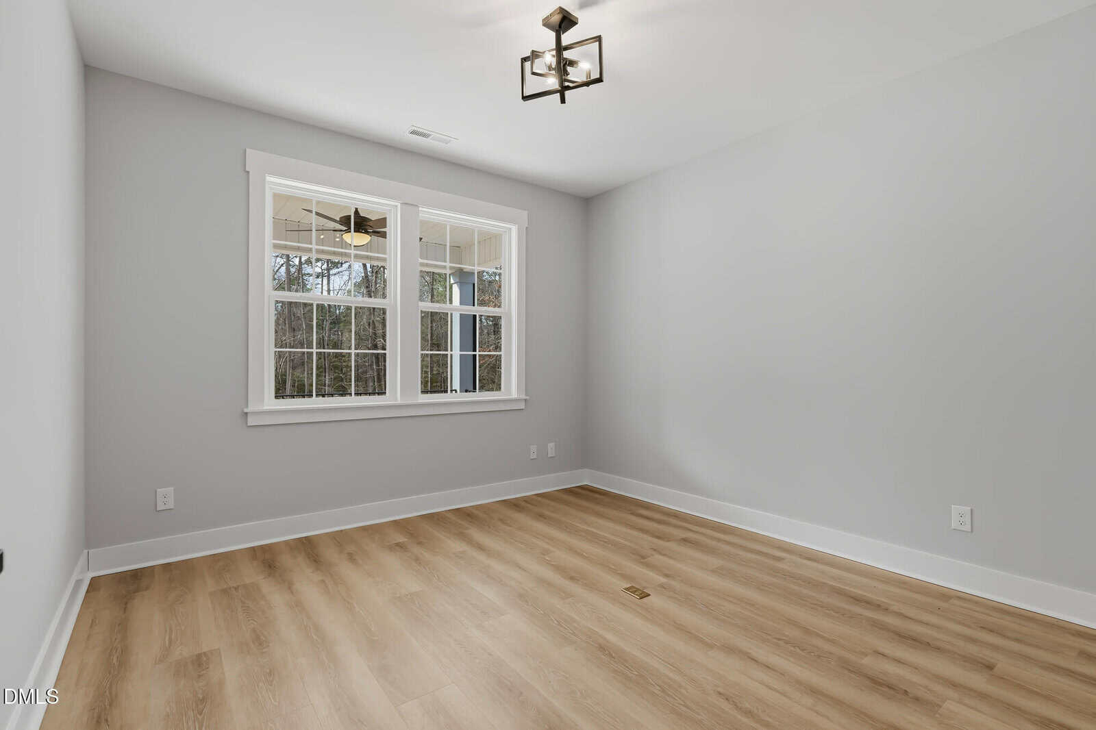 A bright, empty room with light gray walls, light wood flooring, a modern ceiling light fixture, and a large window letting in natural light.