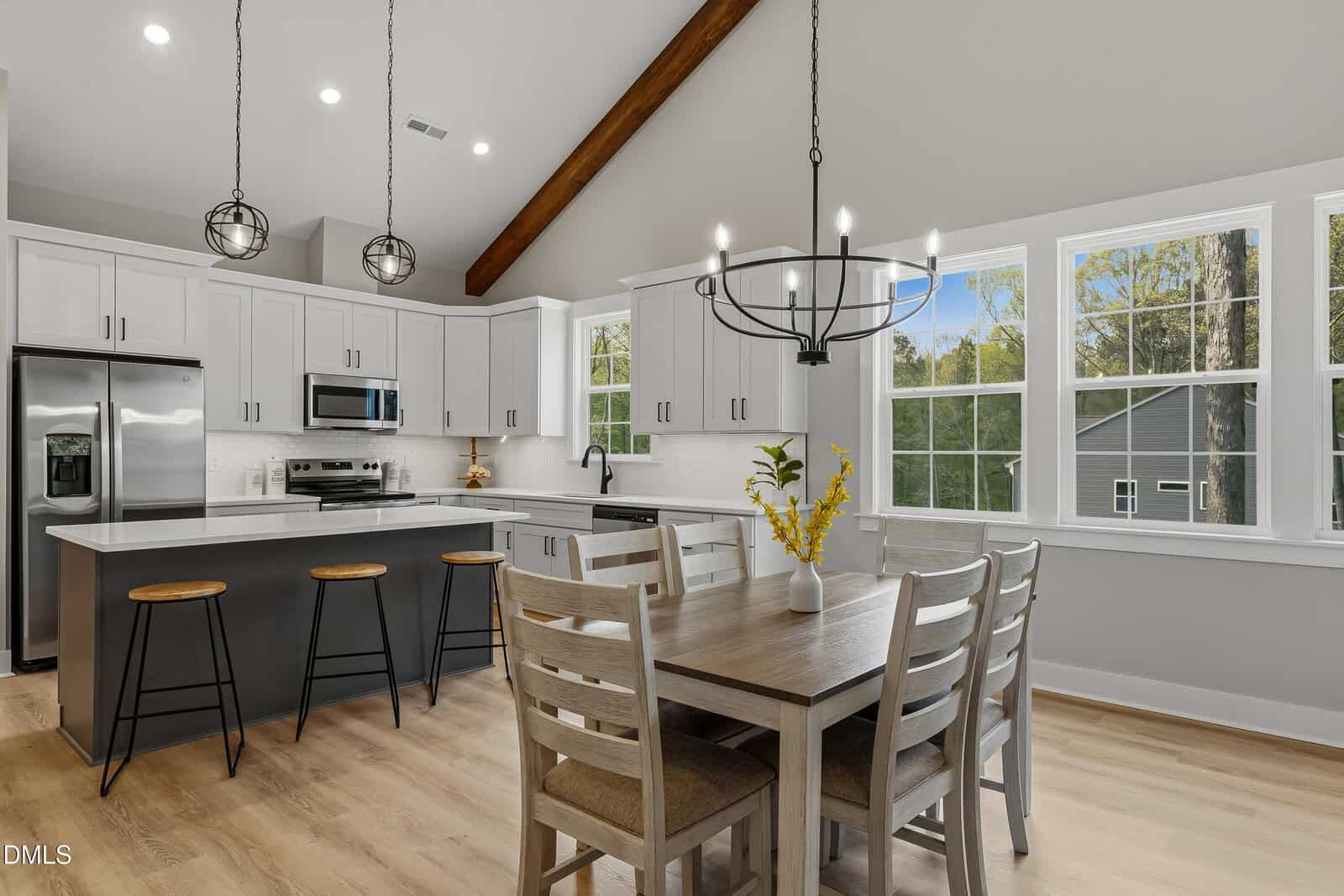 Modern kitchen and dining area with white cabinets, stainless steel appliances, a center island with stools, a wooden dining table with chairs, pendant lights, and large windows letting in natural light.