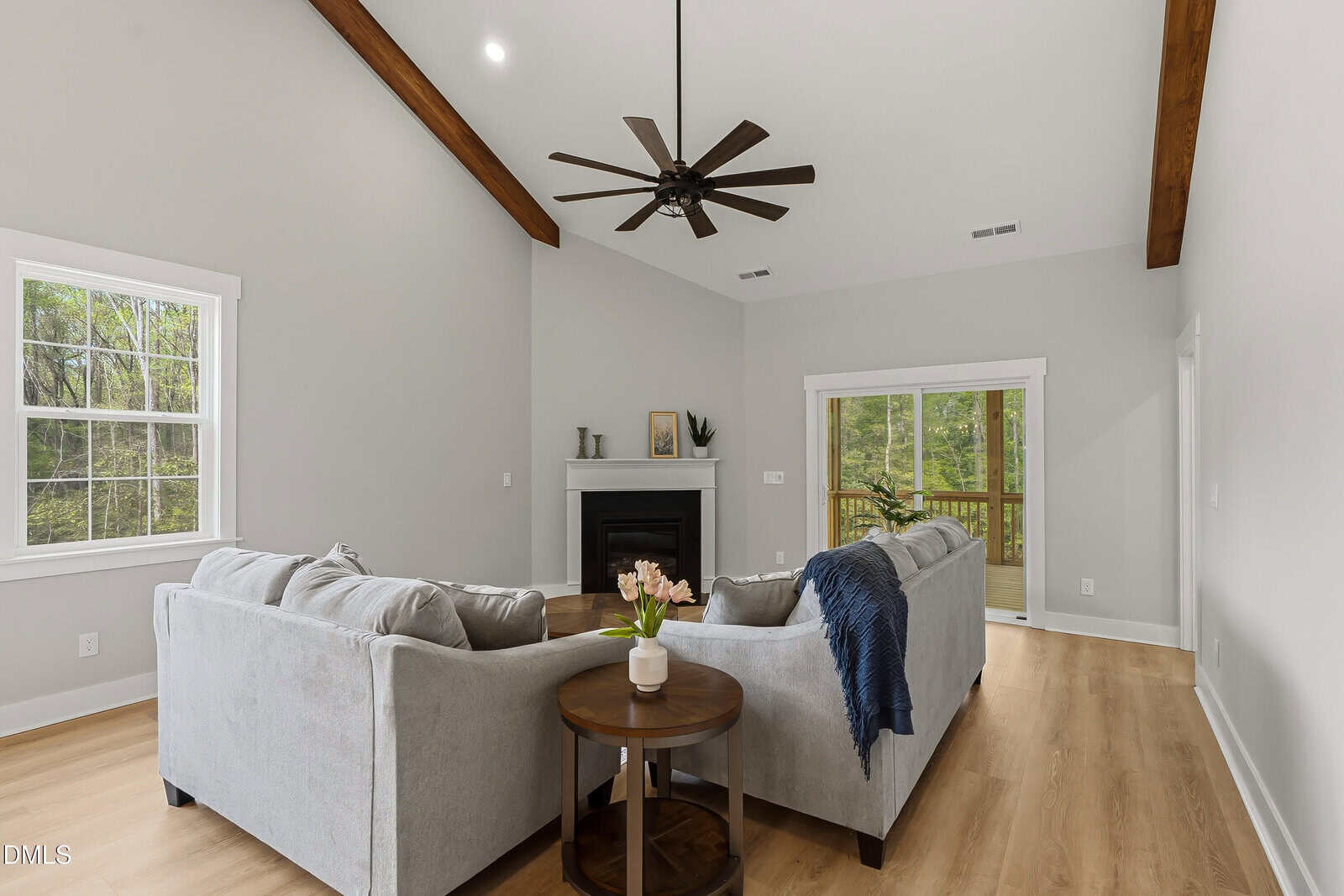 Modern living room with light gray walls, wood floors, two gray sofas, round side table with pink flowers, black ceiling fan, fireplace, and large windows with views of trees outside.