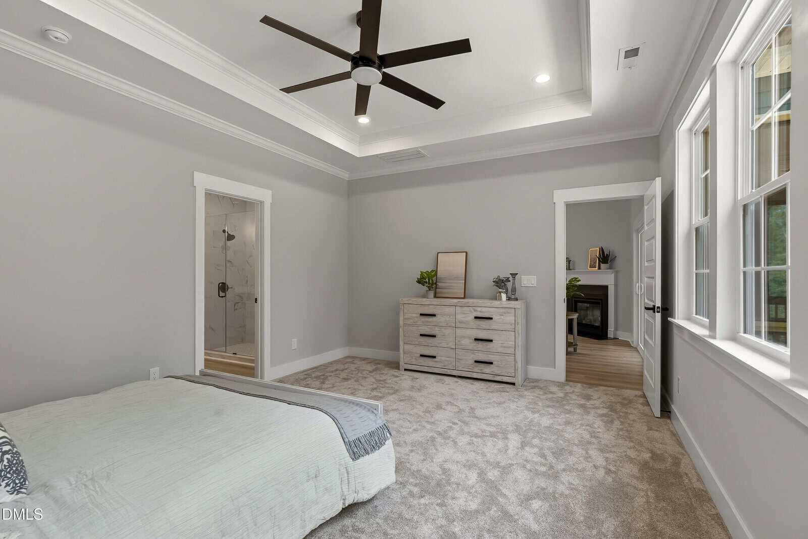 A modern bedroom with gray walls, carpeted floor, a bed in the corner, a dresser with plants and a mirror, a ceiling fan, and open doors leading to a bathroom and another room.