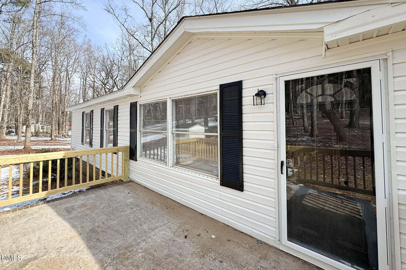 A white single-story house with black shutters, large windows, a sliding glass door, and a wooden ramp on the porch. Leafless trees and patches of snow are visible in the background.