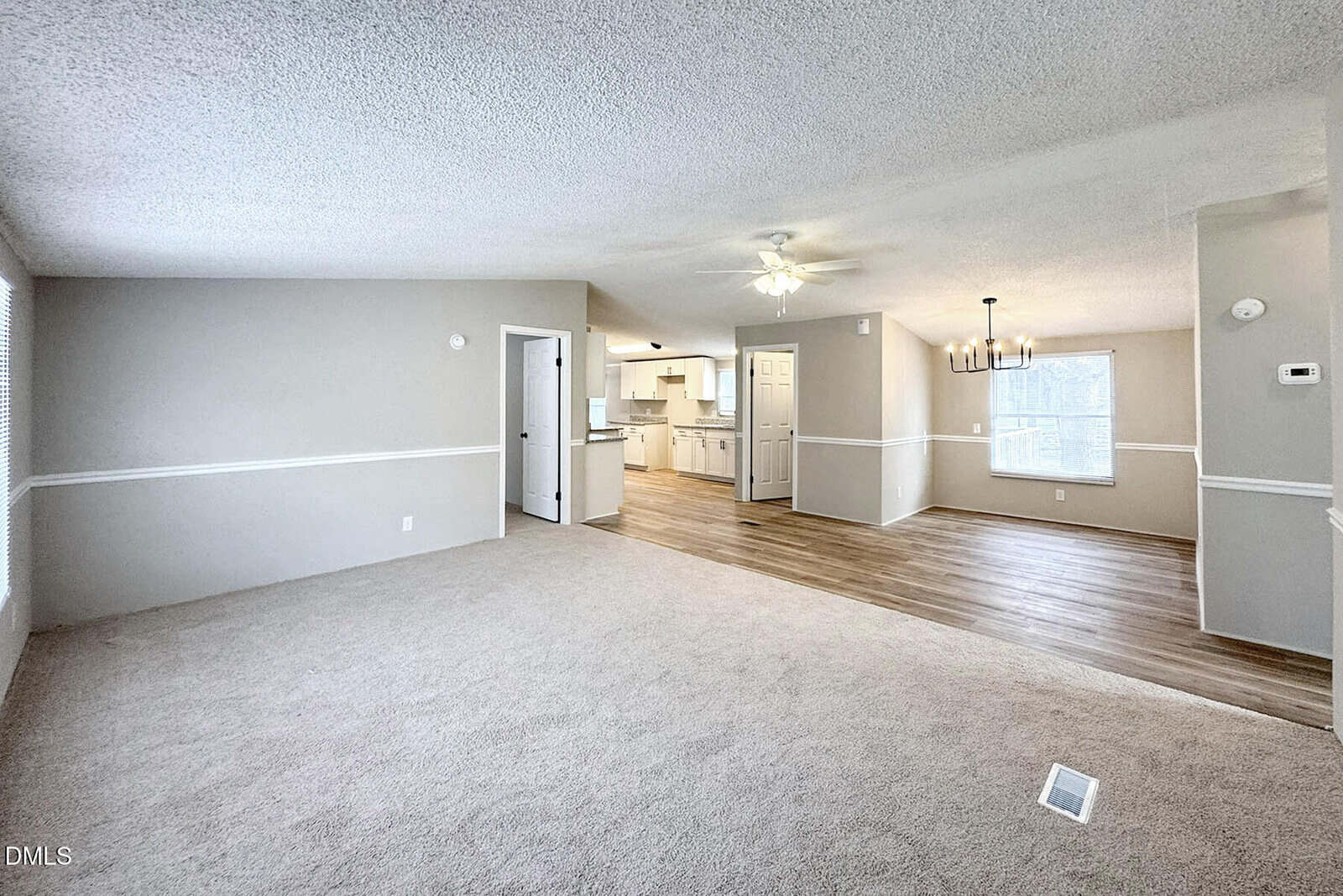 Open living space with beige carpet and light gray walls, leading into a dining area with wood floors. The space features white trim, a ceiling fan, and a view into a bright kitchen with white cabinetry.