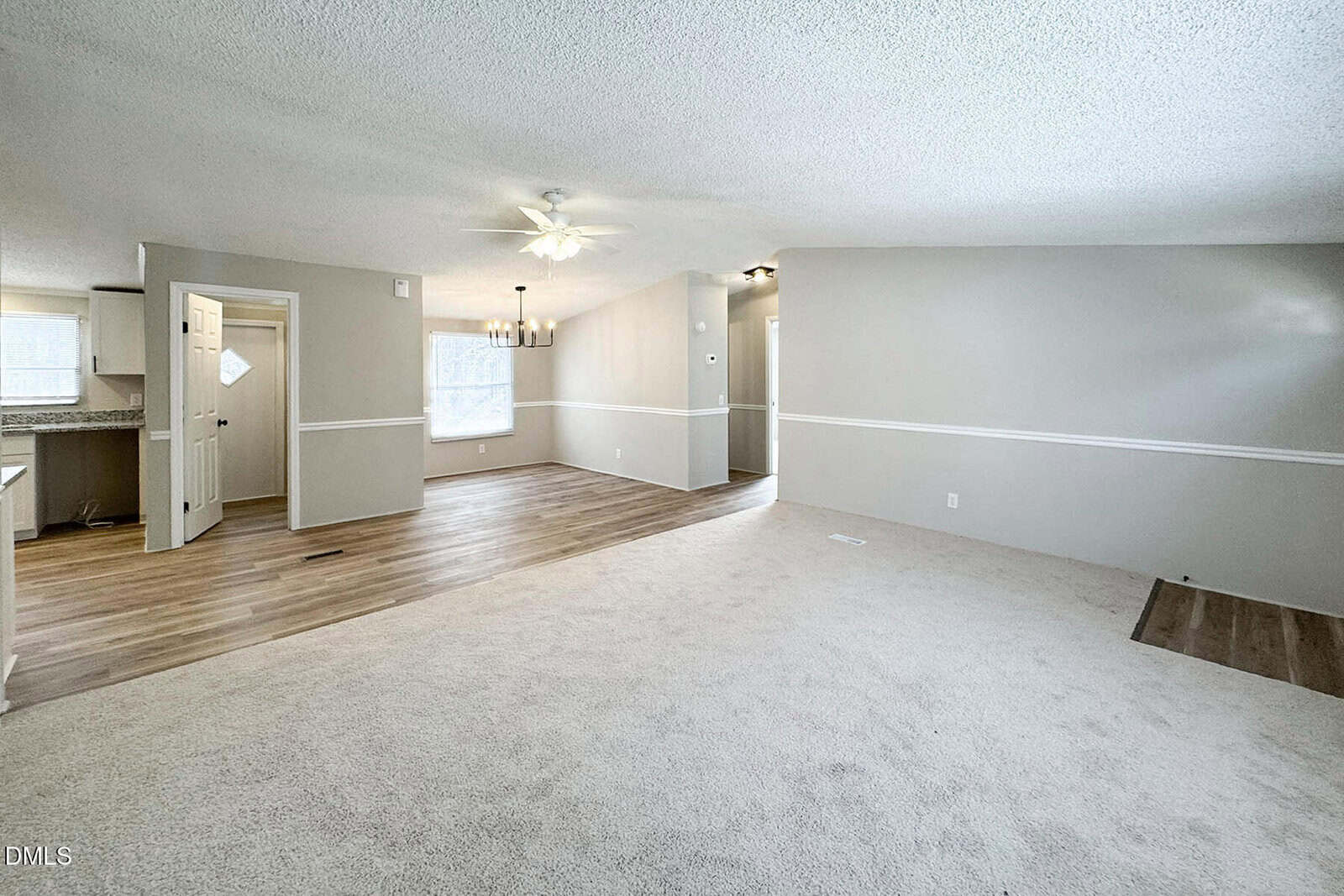 Spacious living and dining area with neutral walls, carpeted floor, and ceiling fan. Dining area has wood flooring, a chandelier, and large windows. Kitchen is partially visible to the left.