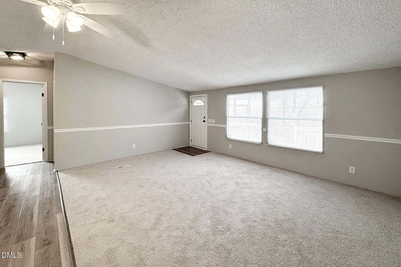A spacious living room with light beige carpet, neutral-colored walls, large windows letting in natural light, a ceiling fan, and an entry door. The room connects to a hallway with wood flooring.