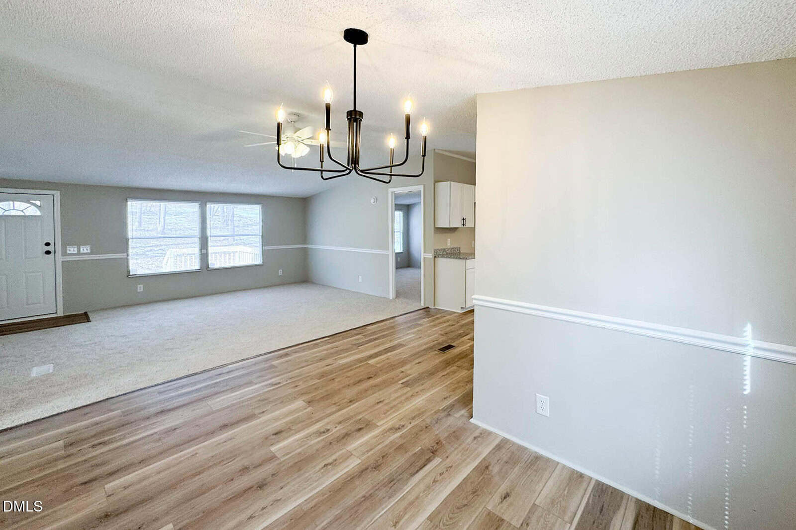 Open, bright living and dining area with light wood flooring, beige carpet, neutral walls, large windows, a modern black chandelier, and a view into a white kitchen.