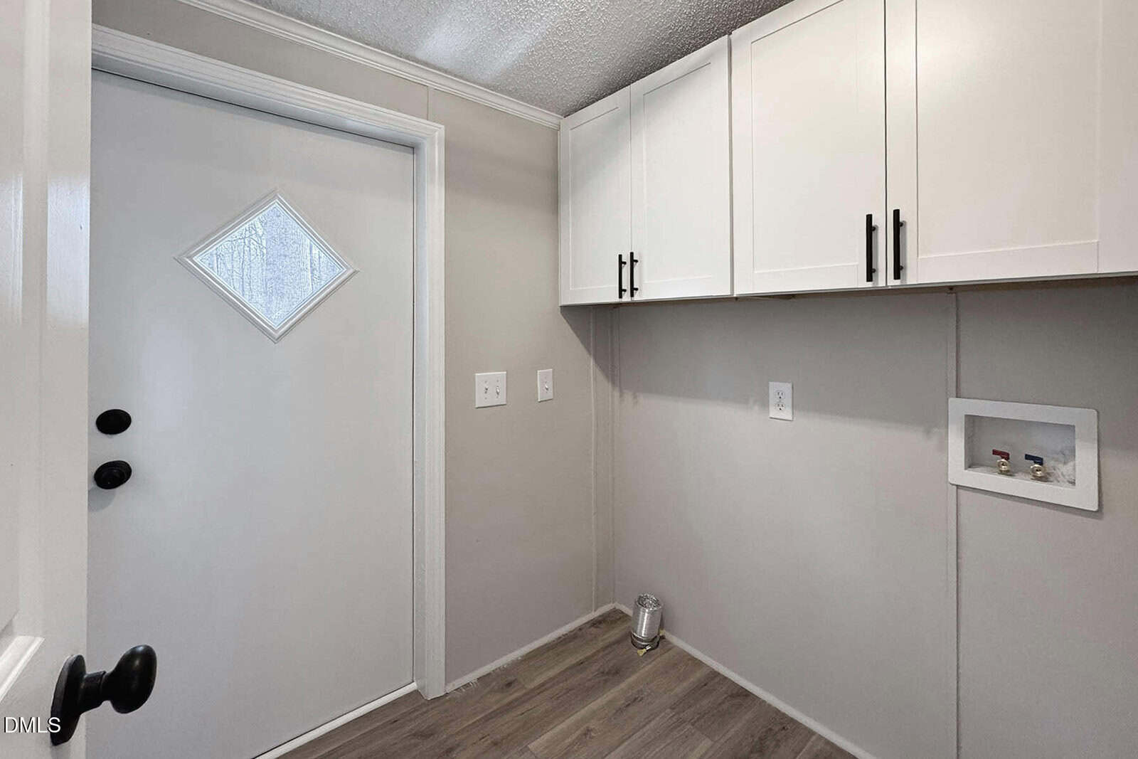A laundry room with light gray walls, white upper cabinets, wood-look flooring, a white door with a diamond window, and washer/dryer hookups on the right wall.