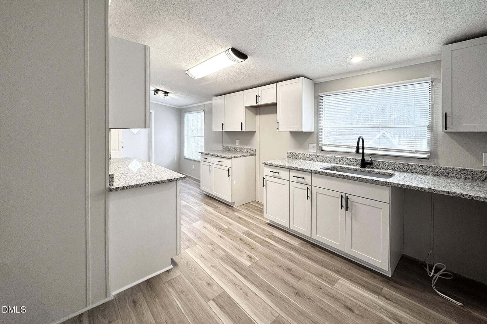 Modern kitchen with white cabinets, granite countertops, stainless sink, and wood-style flooring. Large window above the sink lets in natural light. Open dining area is visible in the background.