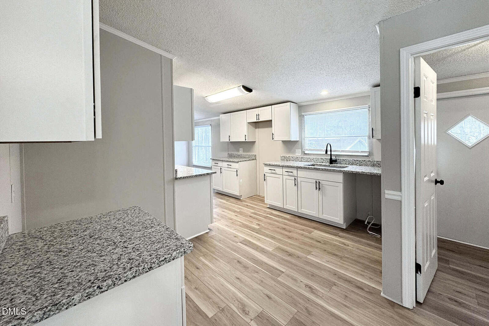 Bright modern kitchen with white cabinets, granite countertops, light wood flooring, and large windows letting in natural light. An open doorway leads to another room.