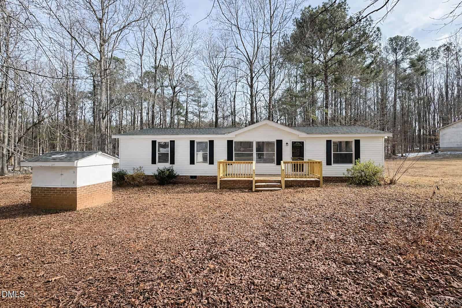 A single-story white house with black shutters sits in front of a wooded area at 2179 Smith Road, Oxford. There is a small wooden porch, light brown mulch covers the yard, and a white shed with a brick foundation is on the left.