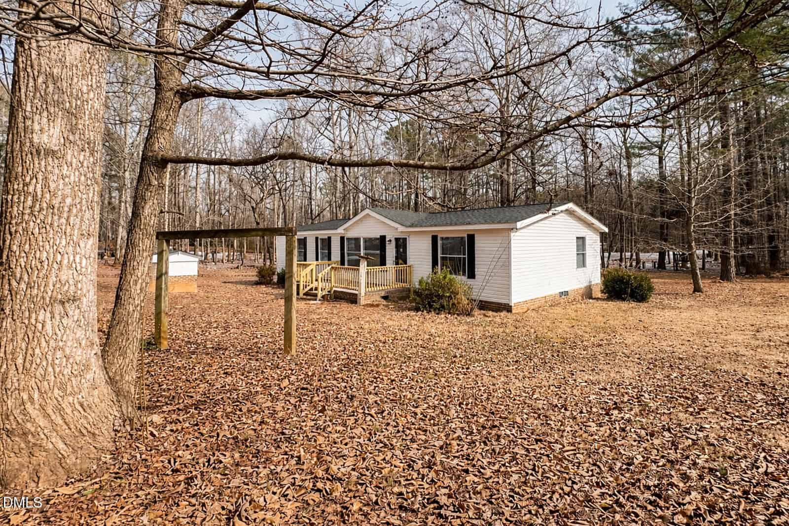 A single-story white house with a front porch sits in a yard covered in fallen leaves, surrounded by tall leafless trees. A wooden frame structure stands near the house. The setting appears quiet and rural.