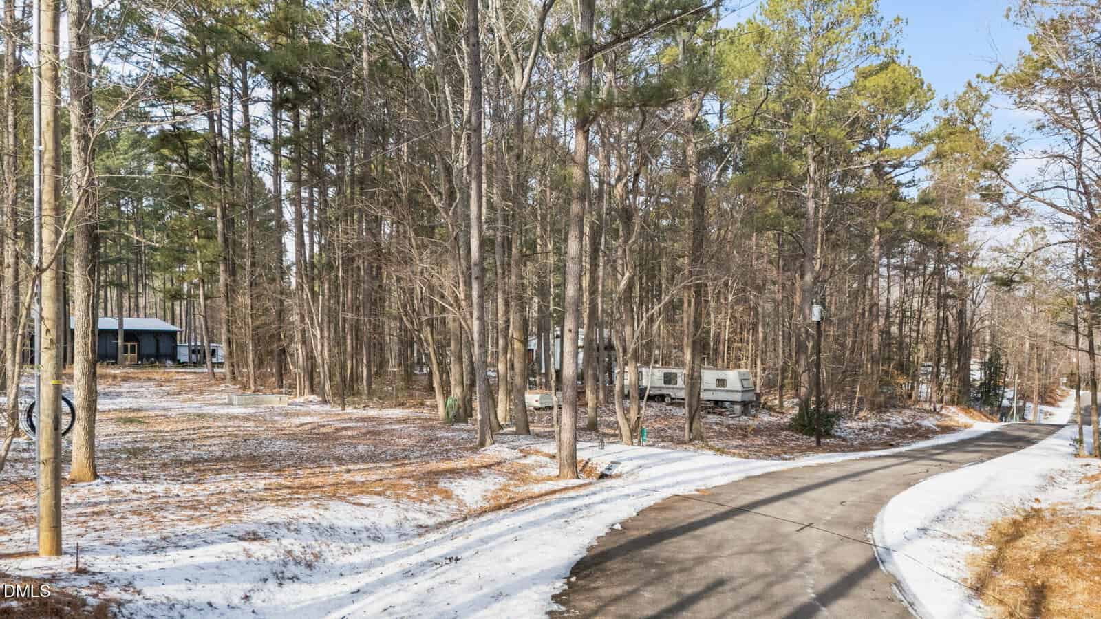 A winding paved road runs through a snowy, wooded area with several RVs and a small cabin visible among tall trees under a clear sky.