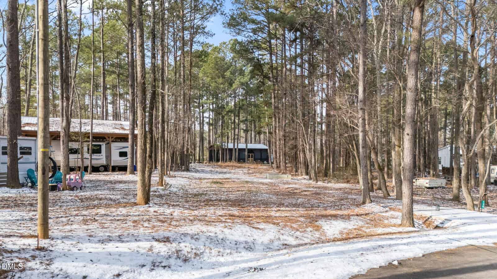 A lightly snow-covered lot in a wooded area with tall trees, two RVs, a swing set, and a small shed visible among the trees under a clear blue sky.