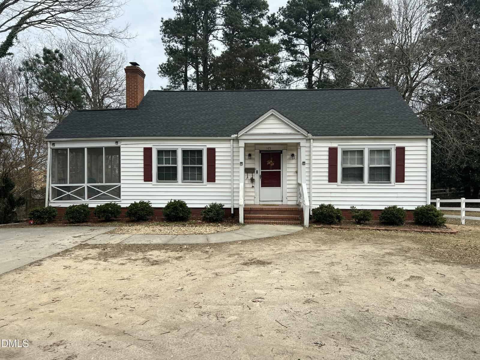 A single-story white house with dark roof, red shutters, and a brick chimney. The front has a small porch, steps, and bushes, with a screened-in side porch. The yard is bare, with trees and a driveway visible.