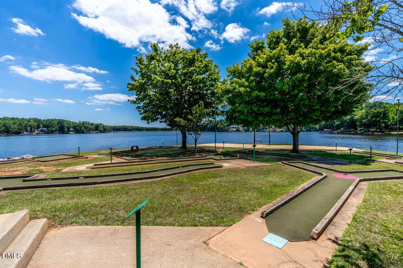 Mini-golf course beside a lake, with green trees providing shade, calm blue water, and a partly cloudy sky in the background.