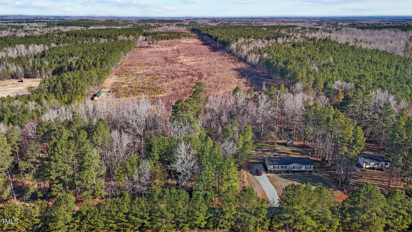 Aerial view of a house surrounded by dense forest with a large, cleared strip of land running through the trees; another house and several vehicles are visible nearby.