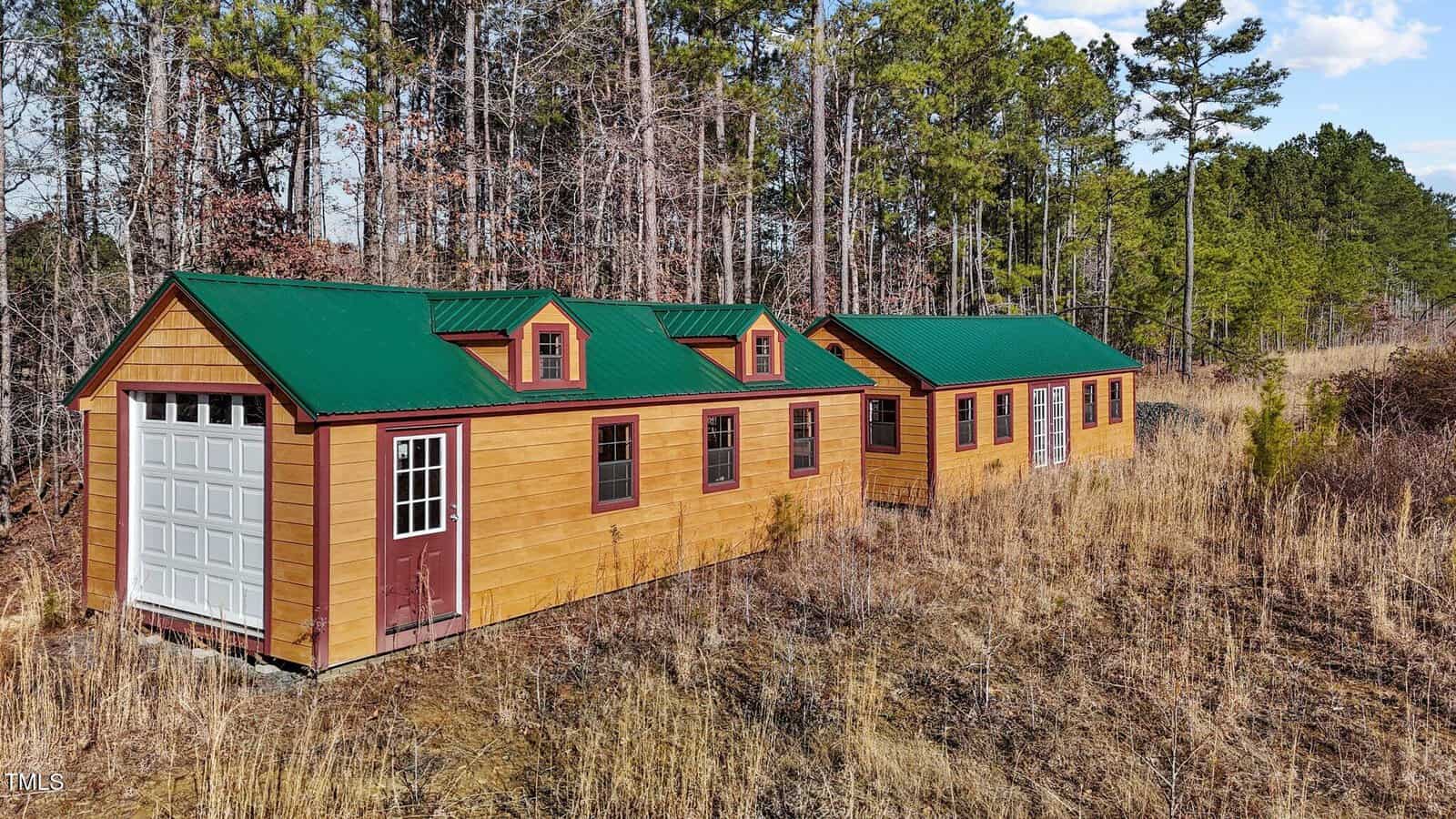 Two small wooden buildings with green metal roofs and red trim sit in a grassy, overgrown clearing near a forest of tall pine trees under a partly cloudy sky. One building has a garage door; the other has double doors and windows.
