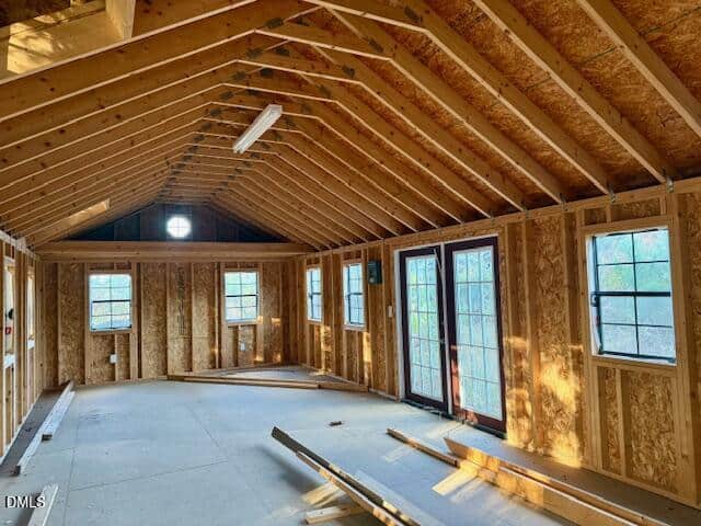 Interior of an unfinished room with exposed wooden framing, a vaulted ceiling, multiple windows, and double glass doors letting in natural light. Construction materials are scattered on the floor.