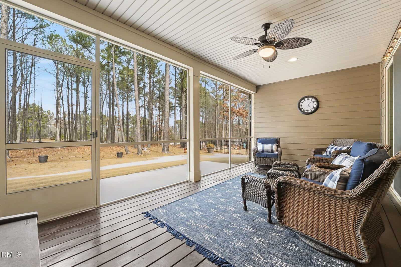 A screened-in porch with wicker seating, blue cushions, a blue rug, a ceiling fan, and a wall clock. Large windows overlook a wooded outdoor area with pine trees and a clear sky.