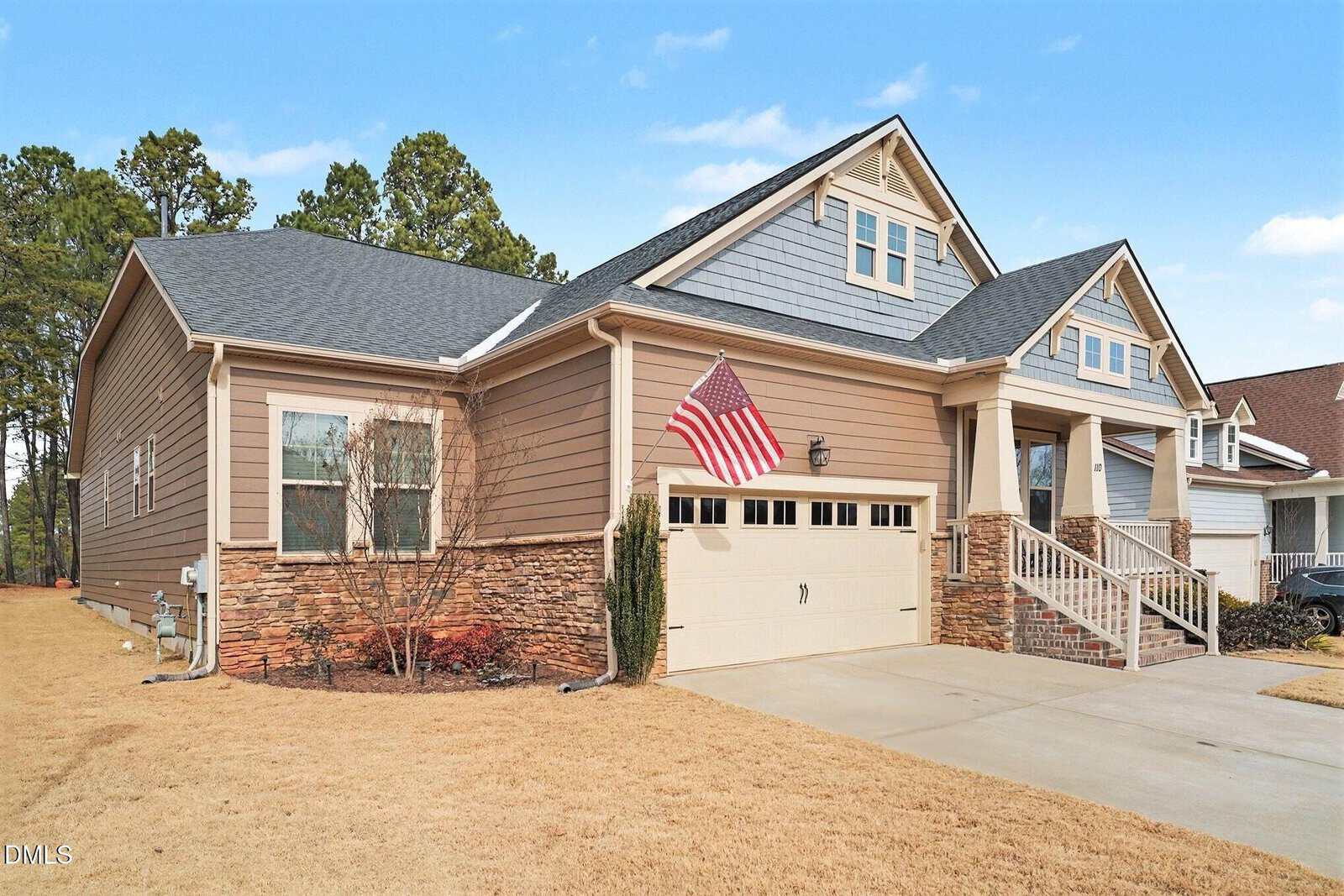A tan two-story suburban house with stone accents, a covered front porch, a two-car garage, an American flag by the entrance, and a dry, yellowish lawn under a clear blue sky.