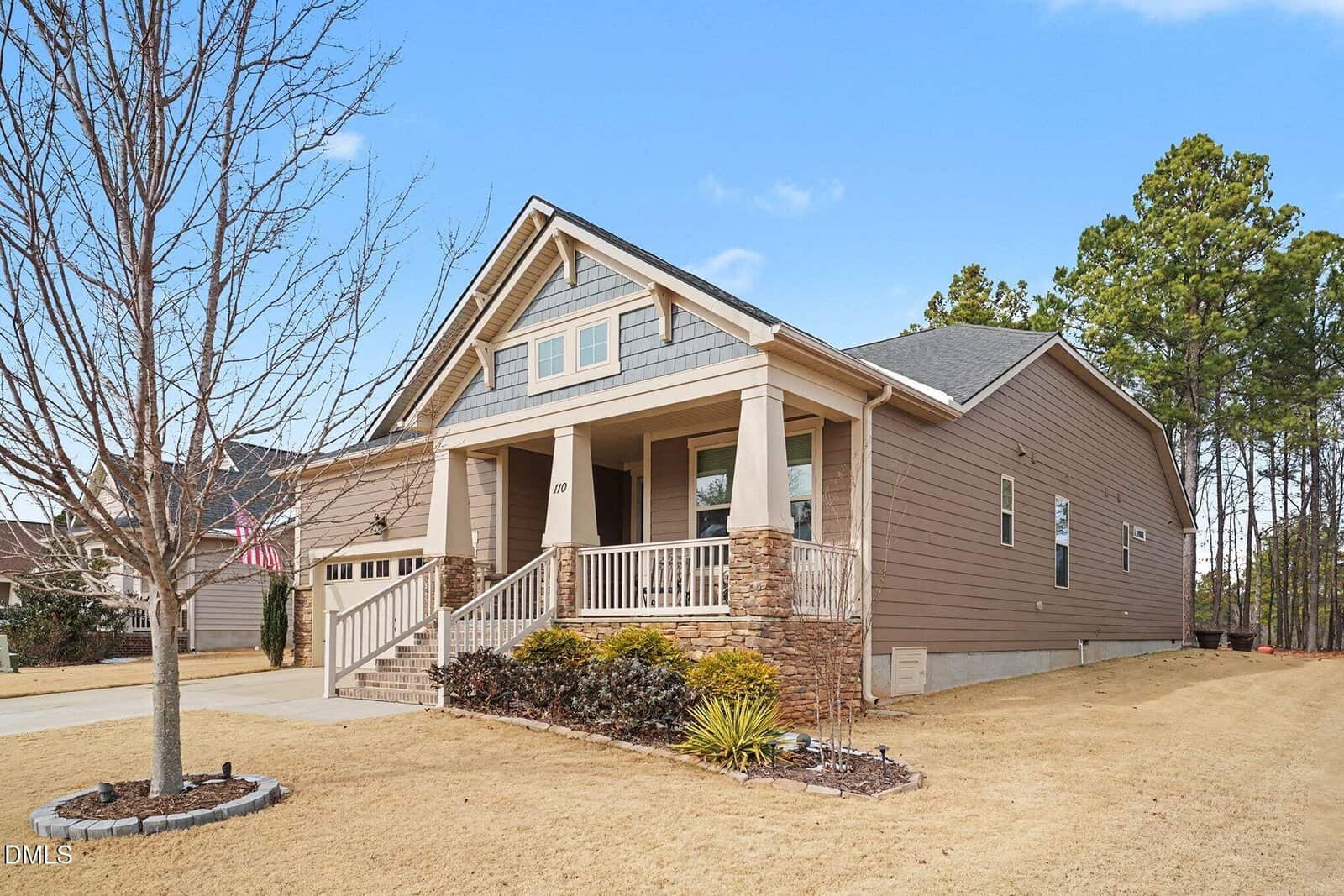 Single-story suburban home with tan siding, stone pillars, a covered front porch, and a two-car garage. The yard has dry grass and minimal landscaping. Leafless trees are in the foreground, with pine trees in the background.
