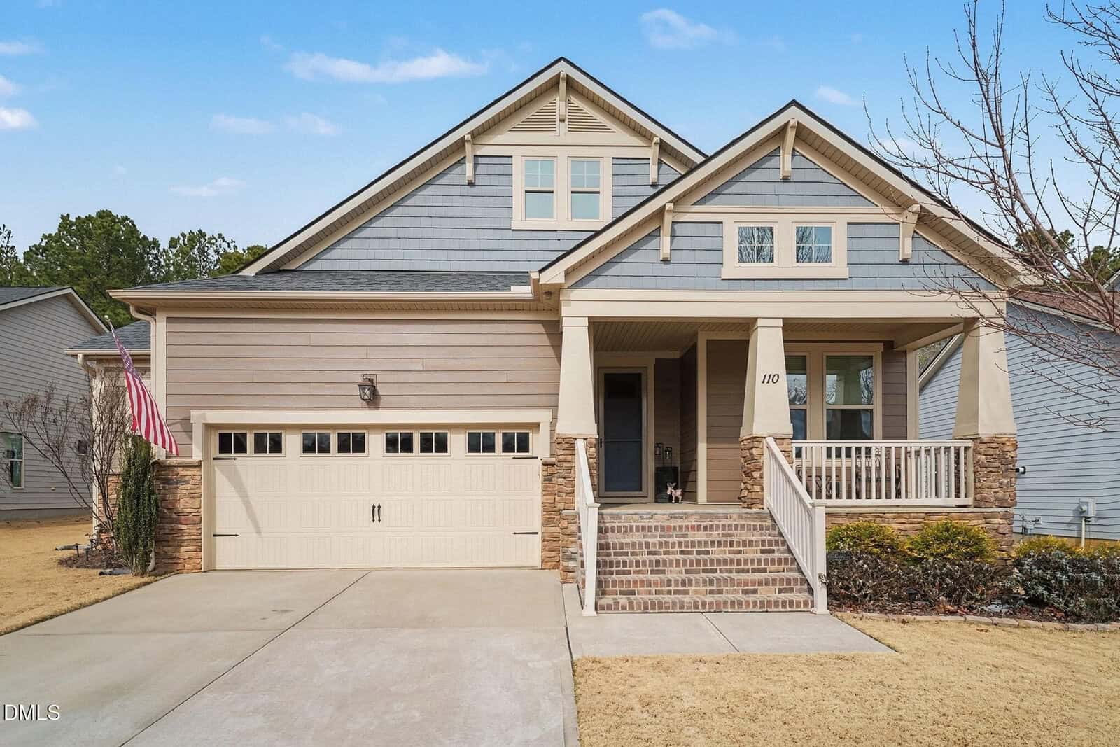 A modern suburban house with gray siding, stone accents, a two-car garage, a front porch with steps, and an American flag by the driveway. The yard has brown grass and light landscaping.