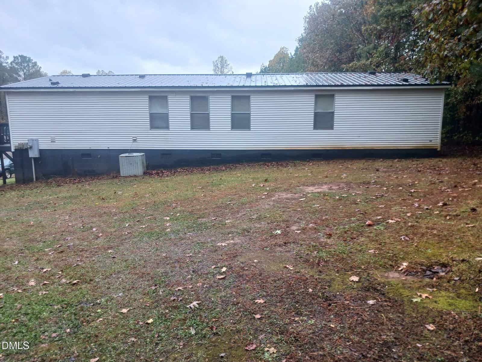 A white manufactured home with a dark foundation and a metal roof sits on a patchy grass yard, surrounded by autumn trees on a cloudy day.