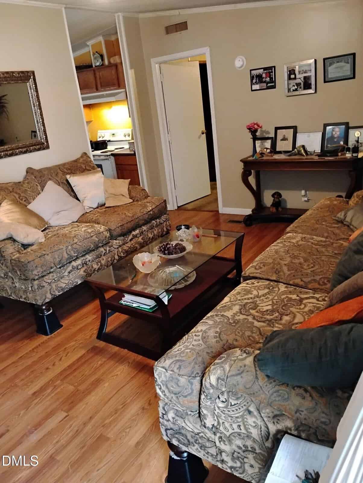 A cozy living room with two patterned sofas, a glass coffee table, wooden floors, and neutral walls. Family photos and decorations are displayed on a console table against the far wall. The kitchen is visible in the background.