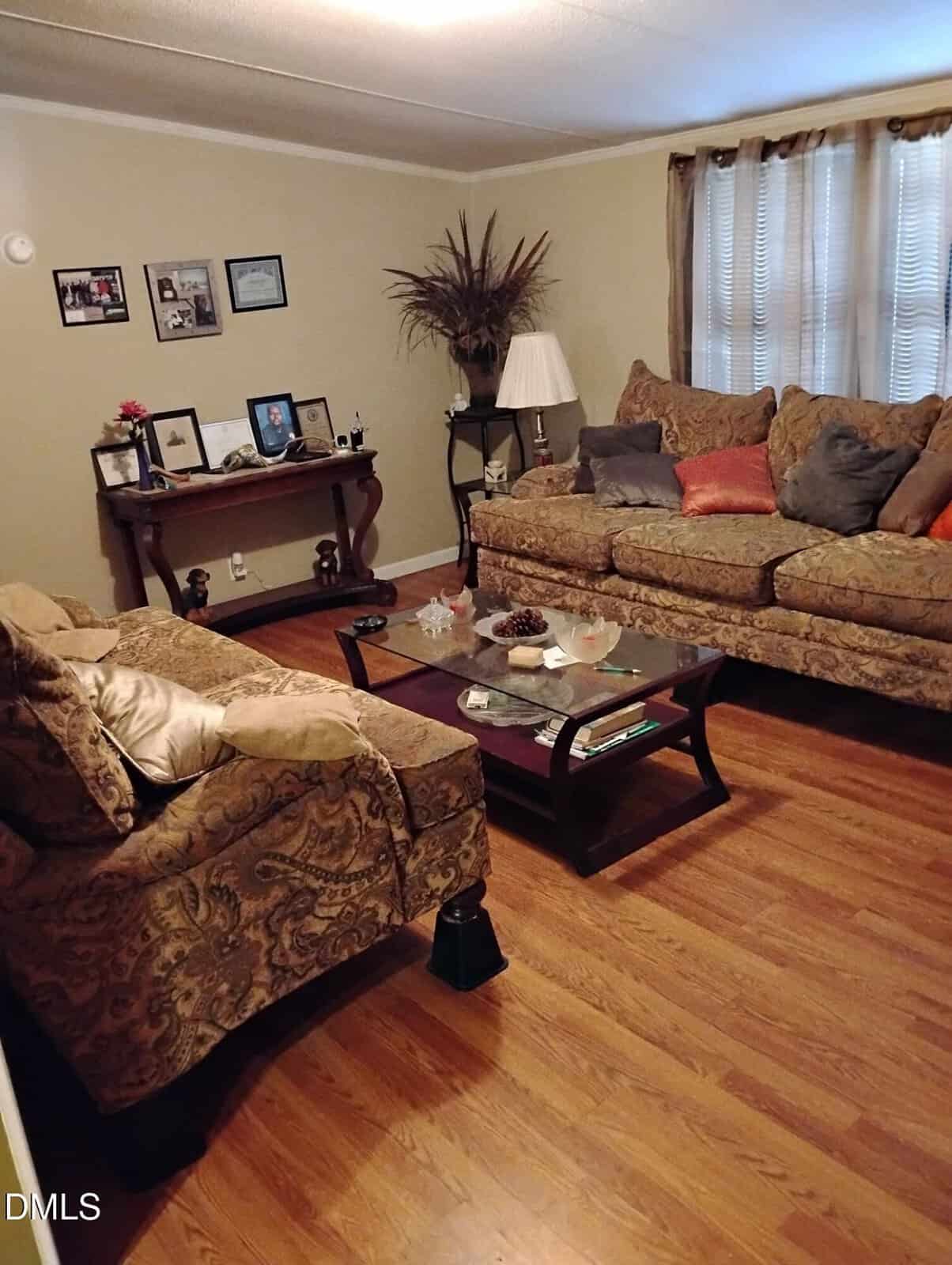 A cozy living room with patterned sofas, a glass coffee table, wood flooring, wall art, framed photos, a lamp, and tall decorative branches in a vase. Light flows in through sheer curtains on the window.