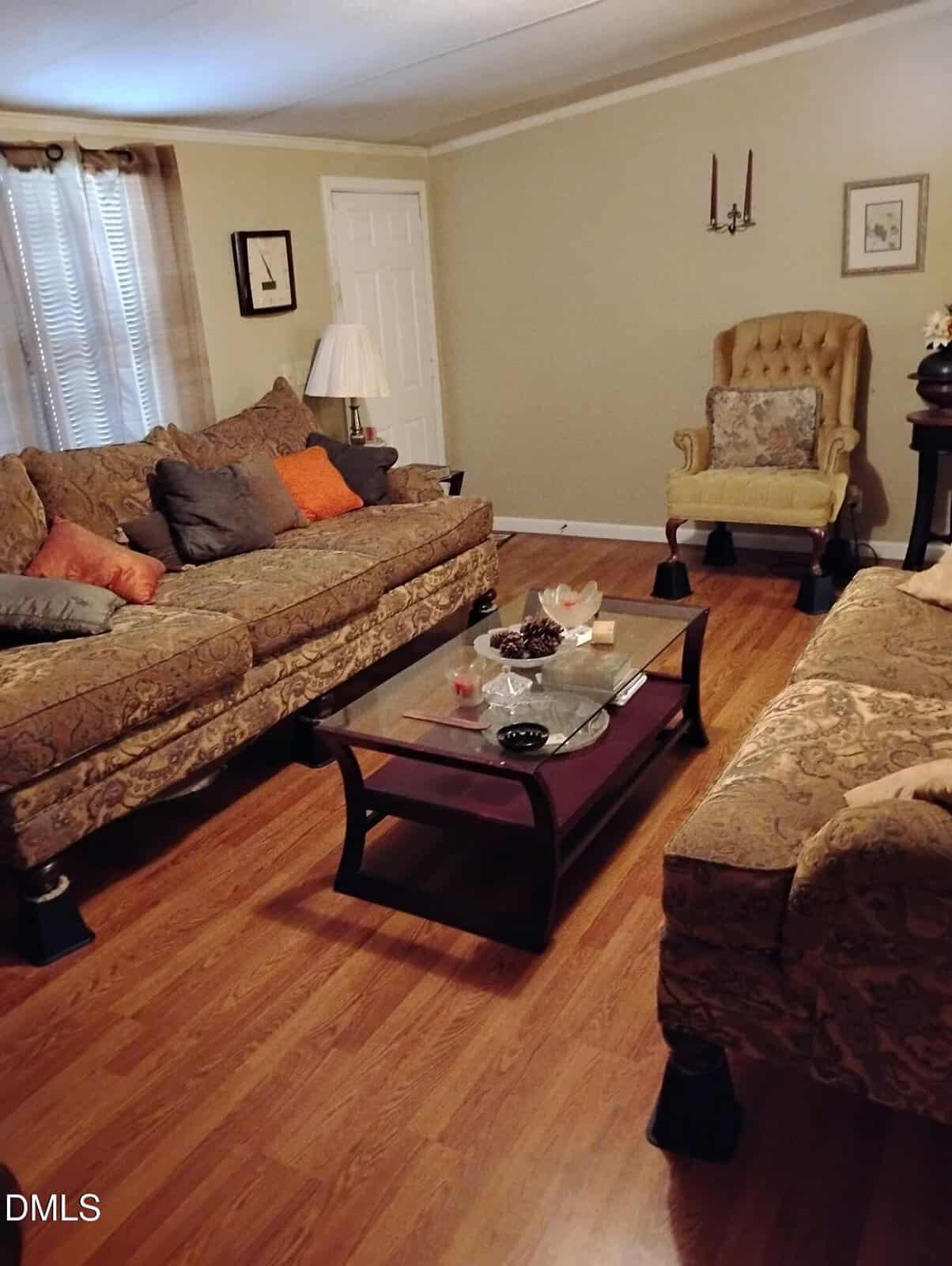A cozy living room with patterned sofas, a glass coffee table, a yellow tufted armchair, hardwood floors, and neutral walls with framed art and a wall sconce. Soft lighting and decorative pillows add warmth.
