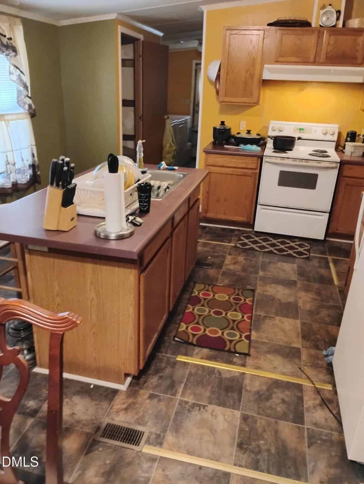 A kitchen with brown cabinets, a central island holding a dish rack and knife block, a white stove and oven, yellow walls, a window with curtains, and patterned rugs on a tiled floor.