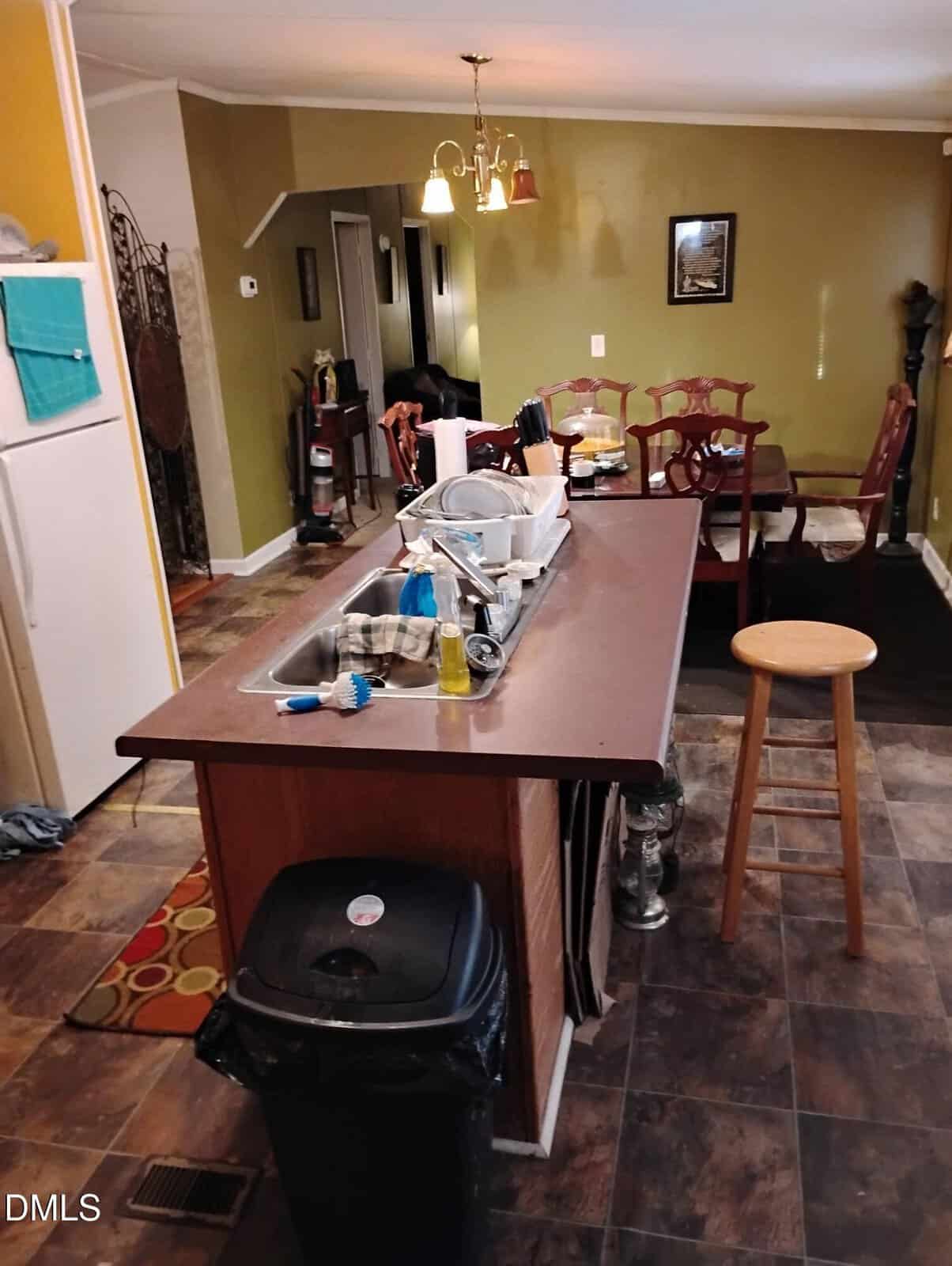 A kitchen with a central island covered in dishes and cleaning supplies, a stool, a trash can, and a view into a dining area with a wooden table and chairs under warm lighting.