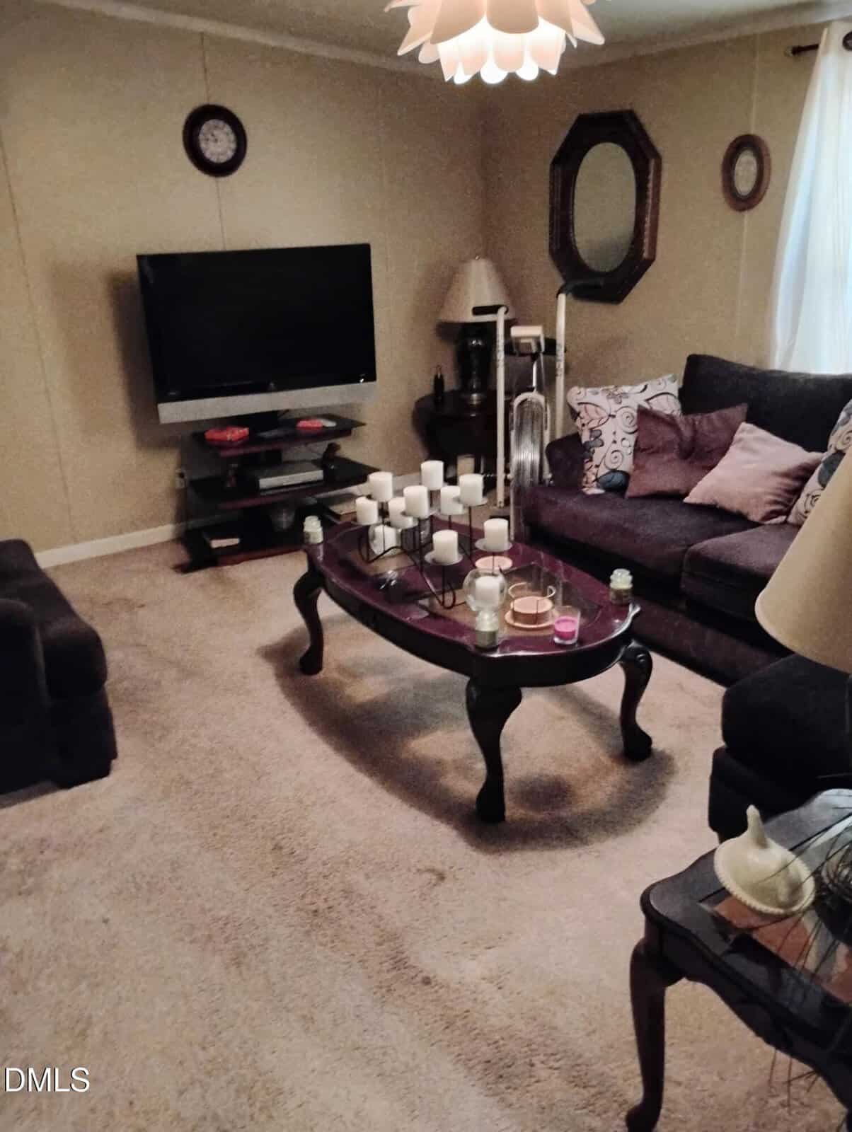 A cozy living room with a sofa, coffee table filled with candles, a wall-mounted TV, a wall clock, mirrors, and end tables with lamps. A window with sheer curtains lets in natural light.
