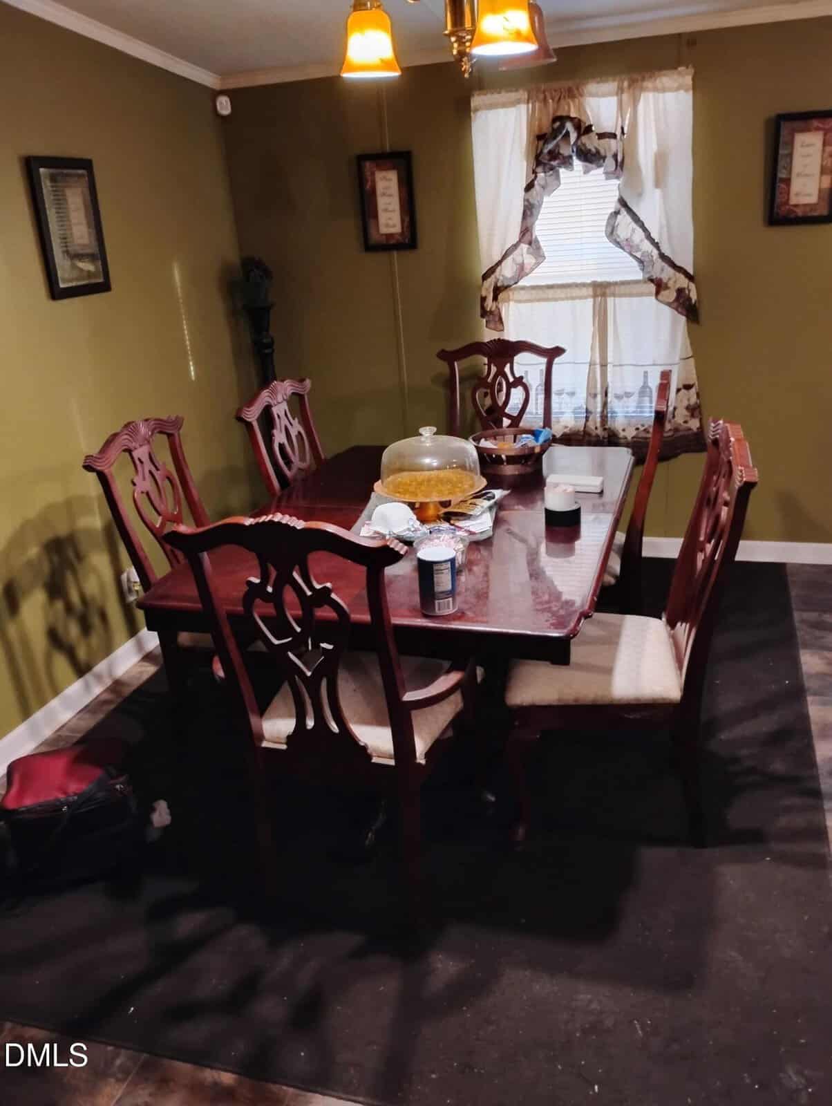 A dining room with a dark wooden table and six matching chairs, a light fixture above, green walls, framed art, a window with patterned curtains, and various items on the table, including dishes and cans.