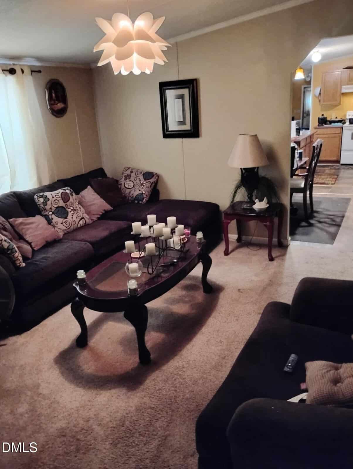 A cozy living room with a sectional sofa and armchair, a glass coffee table with candles, a lamp on a side table, wall art, beige carpet, and a view into a dining area and kitchen in the background.