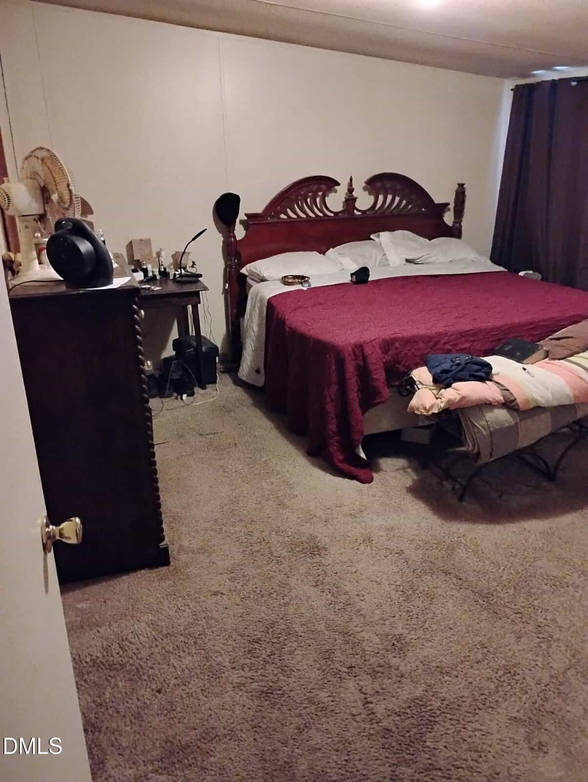 A cozy bedroom with a wooden bed, red bedspread, dresser with mirror, nightstand with various items, and a bench at the foot of the bed. Brown carpet and dark curtains complete the space.