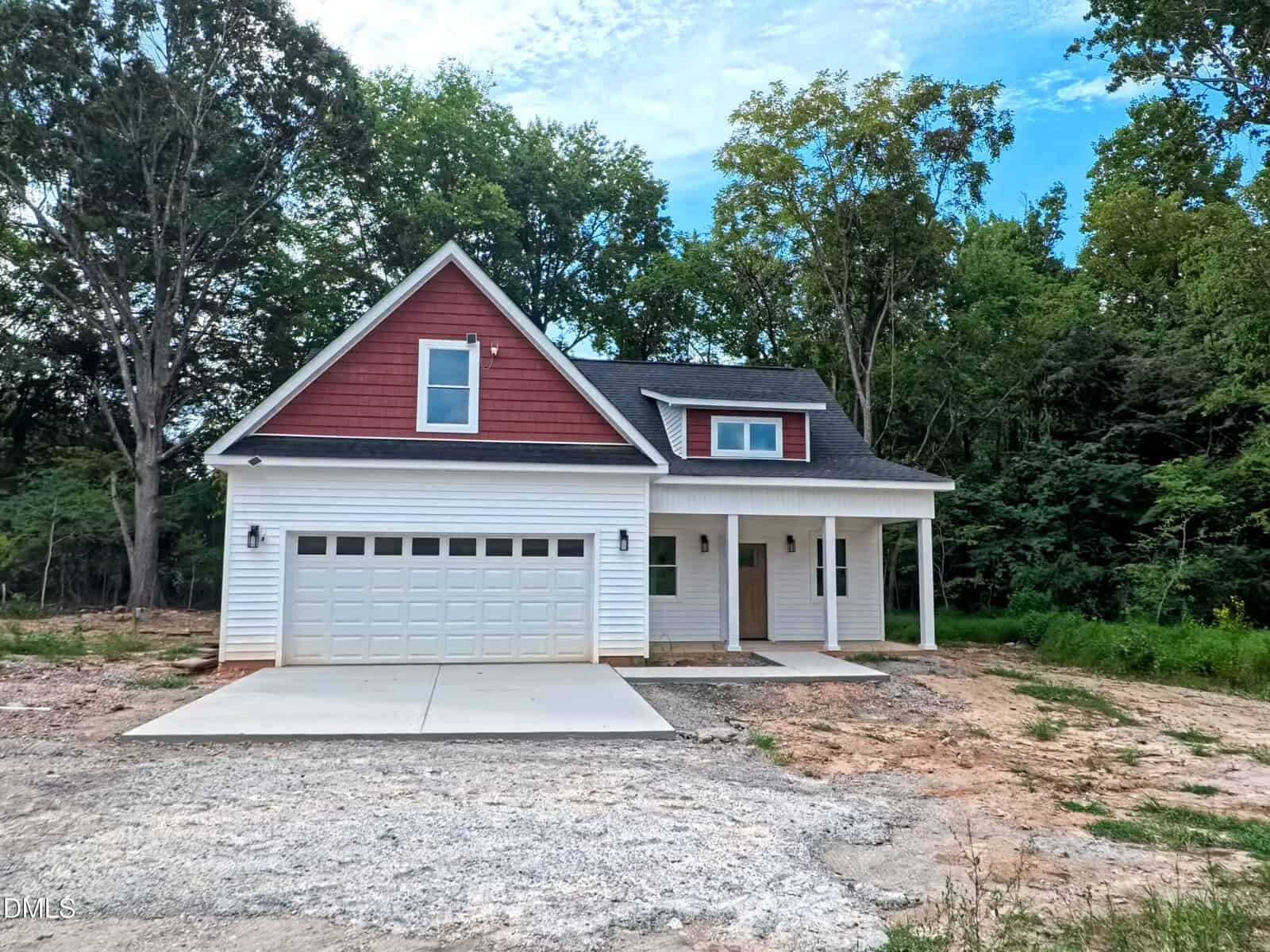 A modern two-story house with a red gable, white siding, a front porch, and a two-car garage sits in front of a wooded area under a partly cloudy sky.